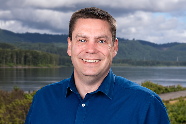 A man with short brown hair, wearing a blue collared shirt, smiles at the camera. Behind him is a lake, green trees, and hills under a cloudy sky.