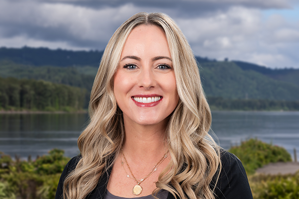A smiling woman with long blonde hair stands outdoors in front of a lake and green hills, under a cloudy sky. She wears a dark top, gold necklace, and a black jacket.
