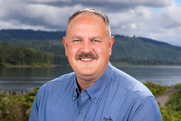 A man with short gray hair and a mustache, wearing a blue collared shirt, smiles while standing outdoors with a lake, trees, and hills in the background under a cloudy sky.